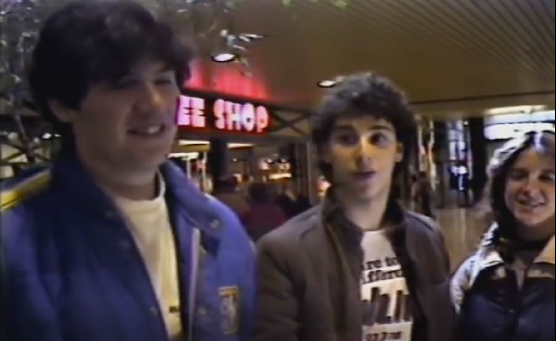 screenshot from the 1983 video documentary "Mall City." From left to right are two young men and one young woman being interviewed by the filmmaker in front of a coffee shop at a Long Island mall.