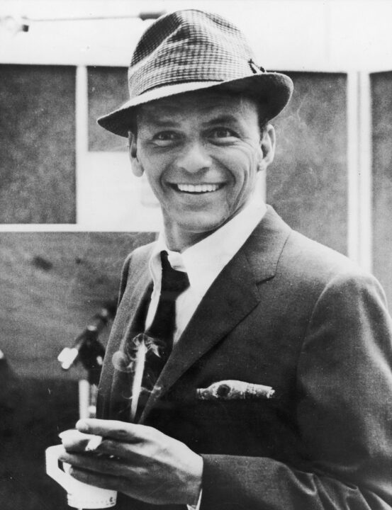 American singer and actor Frank Sinatra (1915 - 1998) smiles while holding a cigarette and a cup of coffee in a recording studio, 1950s.