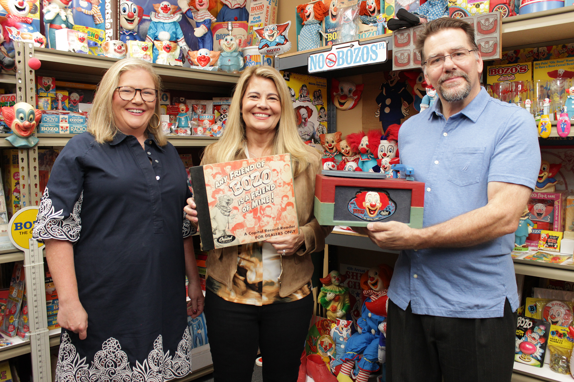 image from a Bozo the Clown-themed episode of MeTV's "Collector's Call." From left to right, each holding Bozo memorabilia, are appraiser Kate Martin, series host Lisa Whelchel and collector Tommy Holbrook