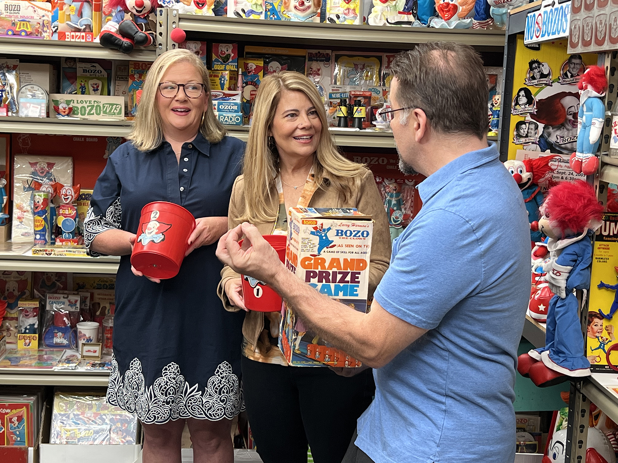 image from a Bozo the Clown-themed episode of MeTV's "Collector's Call." From left to right, standing in a room full of Bozo memorabilia, are appraiser Kate Martin, series host Lisa Whelchel and collector Tommy Holbrook. Holbrook is holding a box of an old toy based on Bozo's "Grand Prize Game," and getting ready to open and play it.