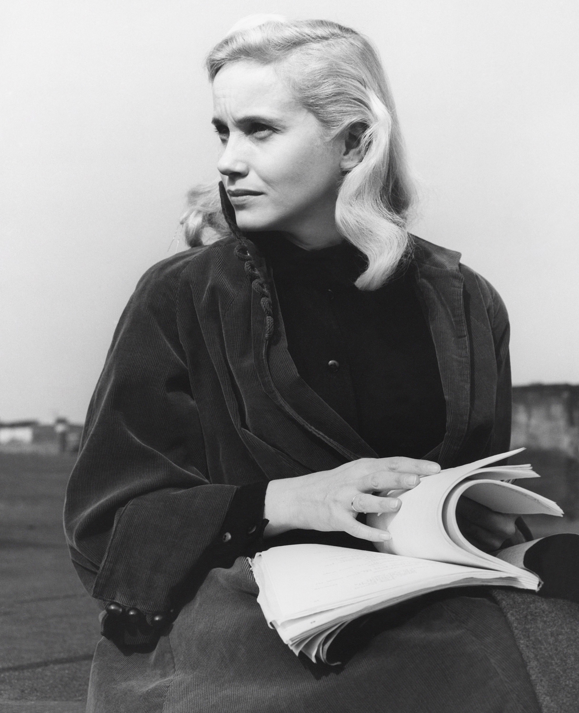 black and white photo from the set of the 1954 movie "On the Waterfront." Star Eva Marie Saint is seated outdoors, wearing a dark coat, and looking up from the script for the film she had been reading.