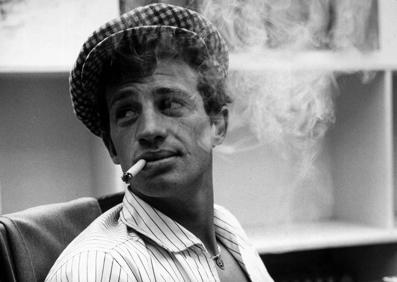 black-and-white image from the 1960 film "Breathless." It is a close-up of star Jean-Paul Belmondo, who is wearing a white shirt and a checkered beret. He is seated and looking to his right with a somewhat bemused toward his right, and holding a cigarette in his mouth. A cloud of smoke from the cigarette is wafting to his left (right in the image).