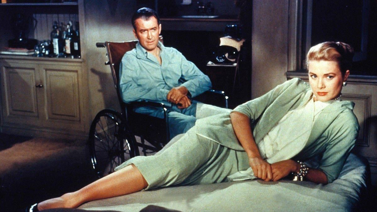 color image from the 1954 film "Rear Window." In the rear background of the shot is James Stewart wearing pajamas and seated in a wheelchair. Spread across the foreground of the image is Grace Kelly, wearing a green dress and lying on her right side on a bed, supported by her left arm, with her right leg crossed over her left. The two are looking at something out of frame.