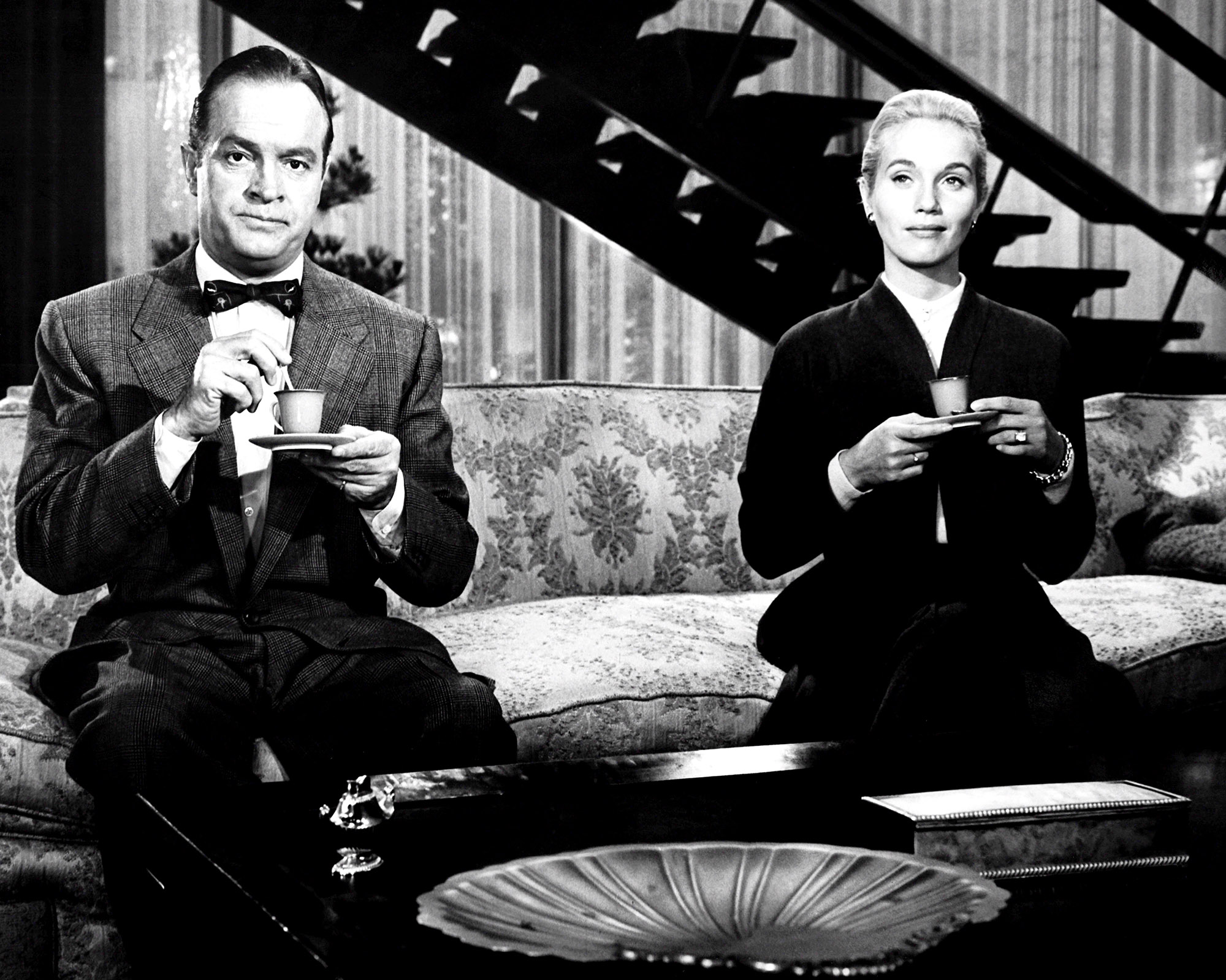 black and white image from the 1956 movie "That Certain Feeling." Star Bob Hope is sitting on one end of a couch, on the left of the picture. On the right of the photo is seated star Eva Marie Saint, with a bit of space between her and Hope. They are both holding cups of tea or coffee on small plates; she has a confident, almost smug look as she looks ahead, while he, also looking ahead, has a more confused look.