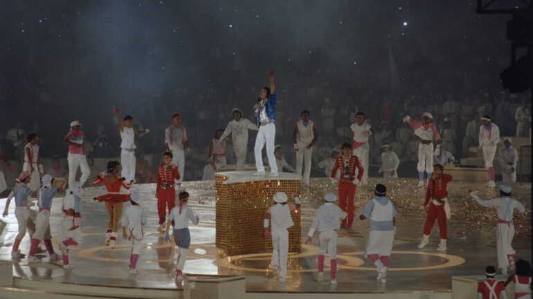 American singer Lionel Richie, wearing a blue jacket and white trousers, on a podium surrounded by performers during the closing ceremony of the 1984 Summer Olympics, held at the Los Angeles Memorial Coliseum in Los Angeles, California, 12th August 1984.