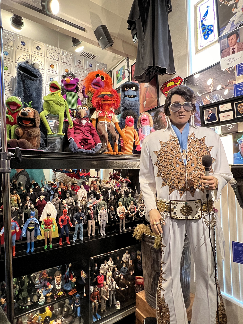 Picture from an episode of the 5th season of "Call from the collector," It features a number of props/costume pieces from Wally Wingert's collection, including a number of Muppets and superhero figures displayed on three levels of a shelf, and an Elvis costume worn by Andy Kaufman.