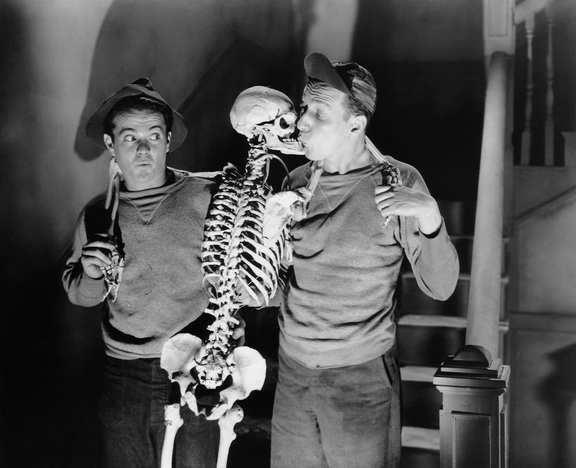 black and white image from the 1943 movie "Ghosts on the Loose." Stars Leo Gorcey, on the left, and Huntz Hall, on the right, have a full standing skeleton between them. The skeleton's head is turned toward Hall, who gives the skeleton a kiss.