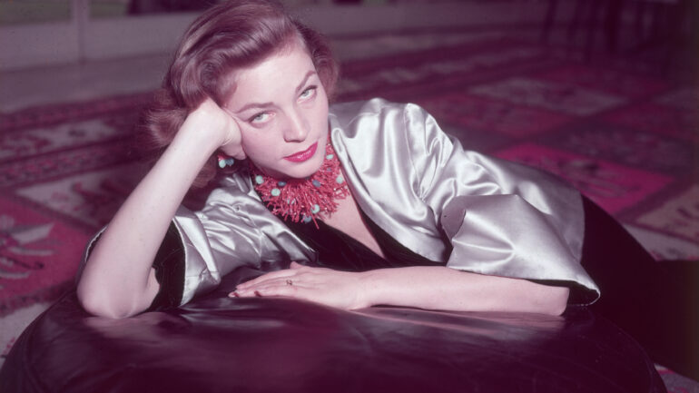 1955: Color portrait of American actor Lauren Bacall wearing a coral necklace and earrings and a gold satin jacket over a black velvet body suit, seated on an oriental carpet and leaning on an ottoman, with the right side of her face cradled in her right hand, bent at the elbow that is rested on the ottoman.