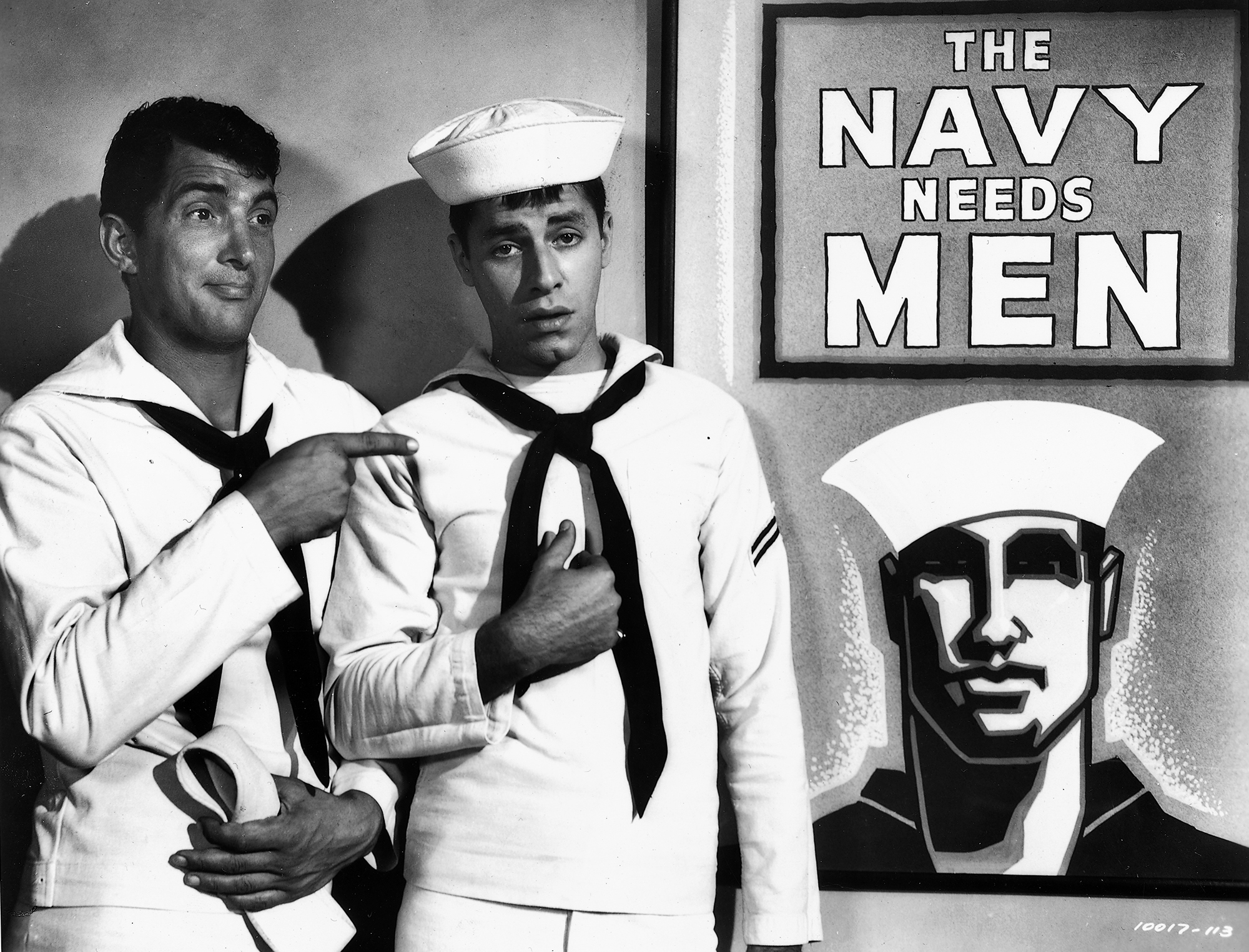 black and white image from the 1952 movie "Sailor Beware." Standing on the left side of the photo, from left to right, are stars Dean Martin and Jerry Lewis. Each is wearing a sailor's uniform, and Lewis also has on his sailor's hat. On the right side of the photo, right to the left of the men, is a poster with an illustration of a sailor, with text above him that reads: "The Navy Needs Men."