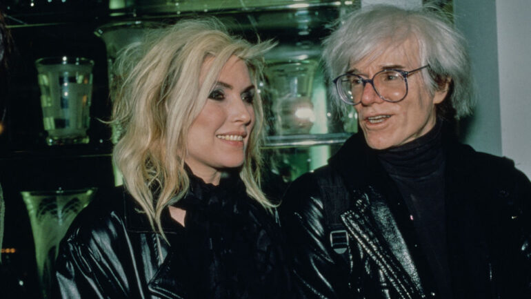 American singer, songwriter and actress Debbie Harry, wearing a black leather jacket and a black scarf, and American artist Andy Warhol, who wears a black leather jacket over a black turtleneck sweater, attend an AIDS Benefit for St Vincent's Hospital, at Barney's in New York City, New York, 10th November 1986