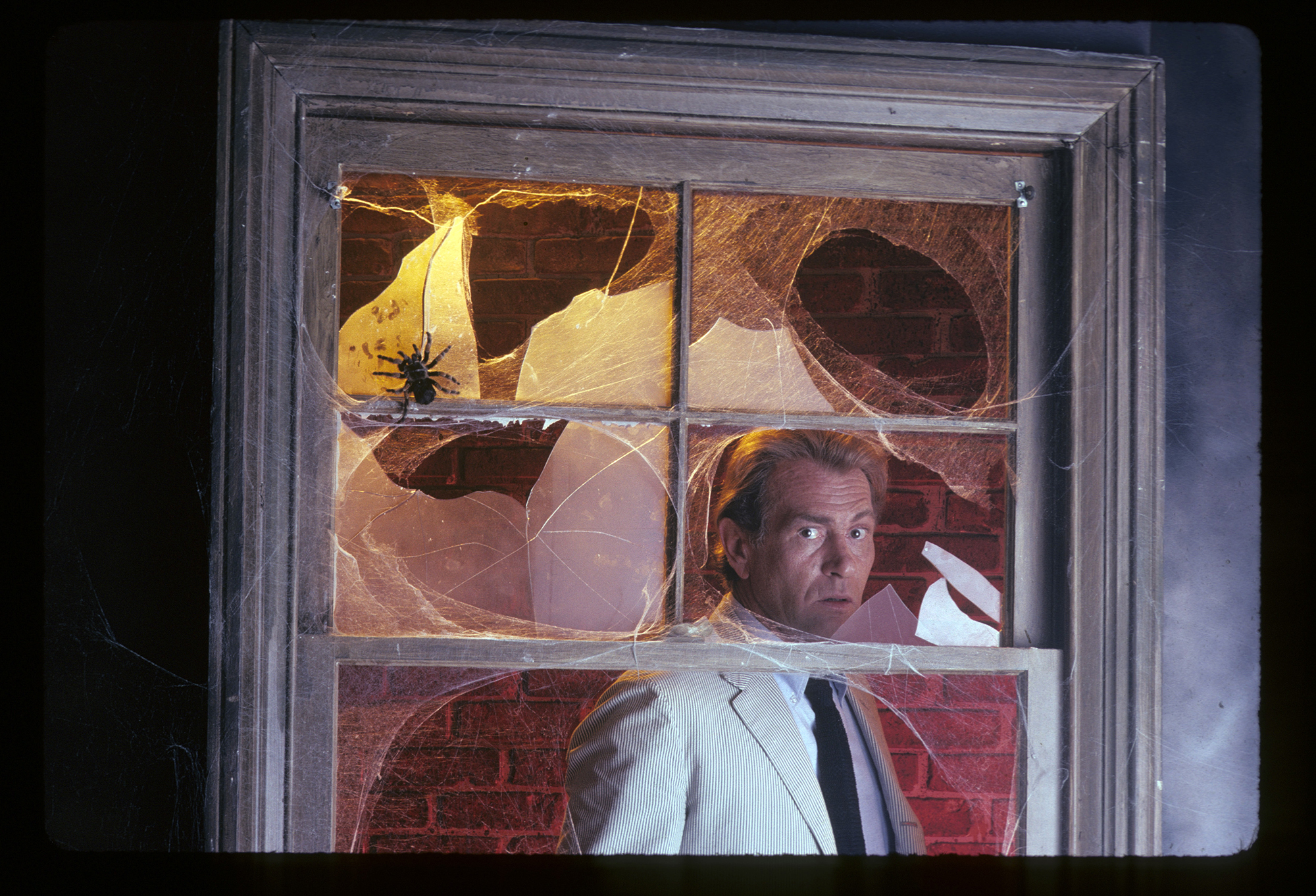 promotional gallery shot for the 1974-75 TV series "Kolchak: The Night Stalker." Star Darren McGavin, as Kolchak, wearing a light-colored suit, black tie and straw fedora hat, is behind the window of a creepy old house. Five of the window's six panes of glass are broken; on the unbroken one, a tarantula is crawling. Kolchak is looking out with alarm.