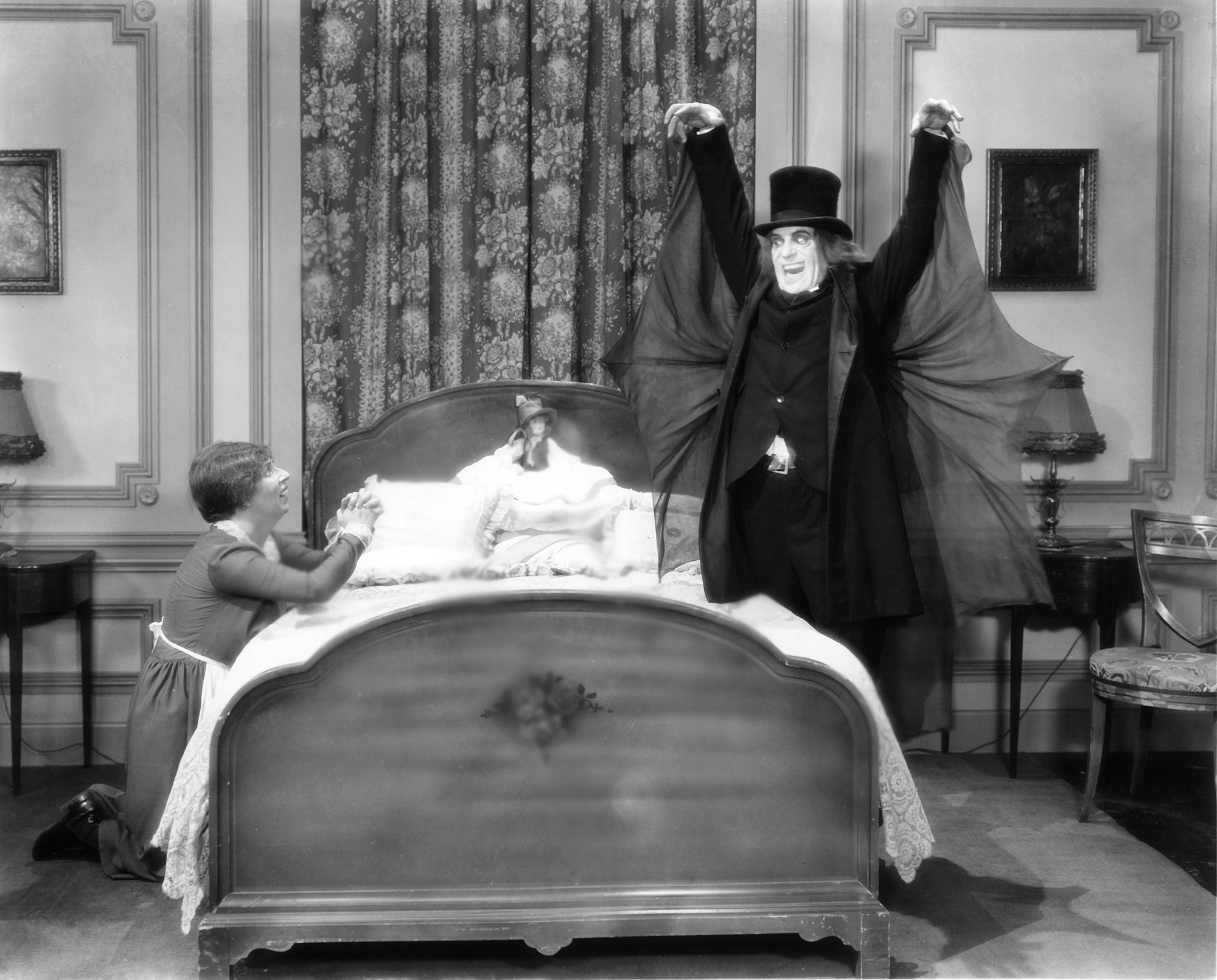 image from the 1927 silent movie "London After Midnight." It is a scene in a bedroom; on the left on the bed, on her knees praying and looking up in fear, is Polly Moran as a maid. She is looking at the other side of the bed, where Lon Chaney's character, wearing a black suit with batlike wings under the arms and a black top hat, with his makeup including sharpened teeth, smiles back at her.