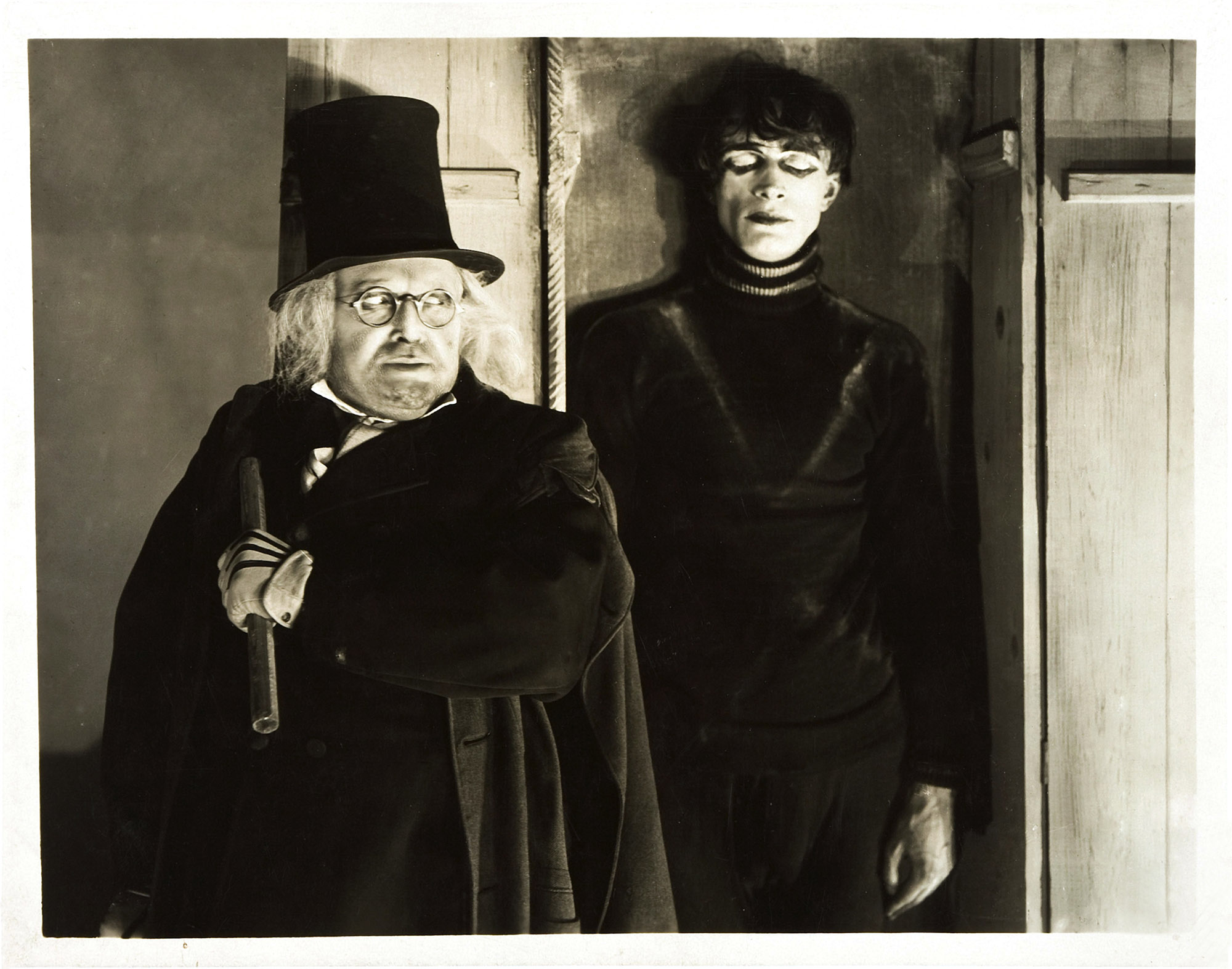 image from the 1920 silent movie "The Cabinet of Dr. Caligari." Standing on the left, wearing a top hat and glasses, is Werner Krauss as Dr. Caligari. Just behind him, and slightly to the right, standing in the cabinet with his eyes closed, is Conrad Veidt as Caligari's brainwashed subject Cesare.