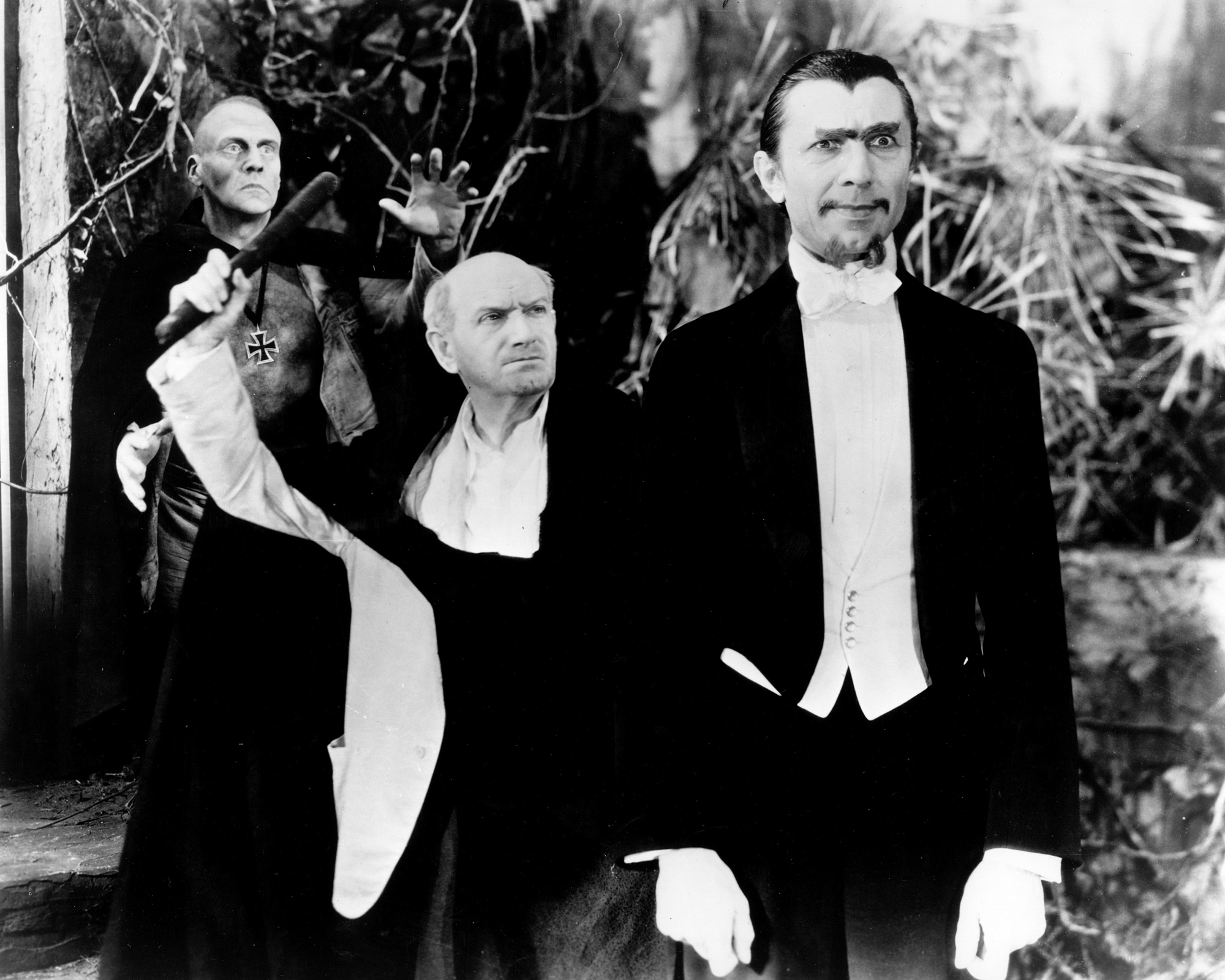 photo from the 1932 movie "White Zombie." Standing on the right is star Bela Lugosi, wearing a dark suit jacket and pants over a white formal shirt and bowtie. He has large, bushy eyebrows, a mustache just on the ends of the lip (shaved in the middle) and a similar type of goatee. He has an evil hypnotic stare as he gazes in front of him.