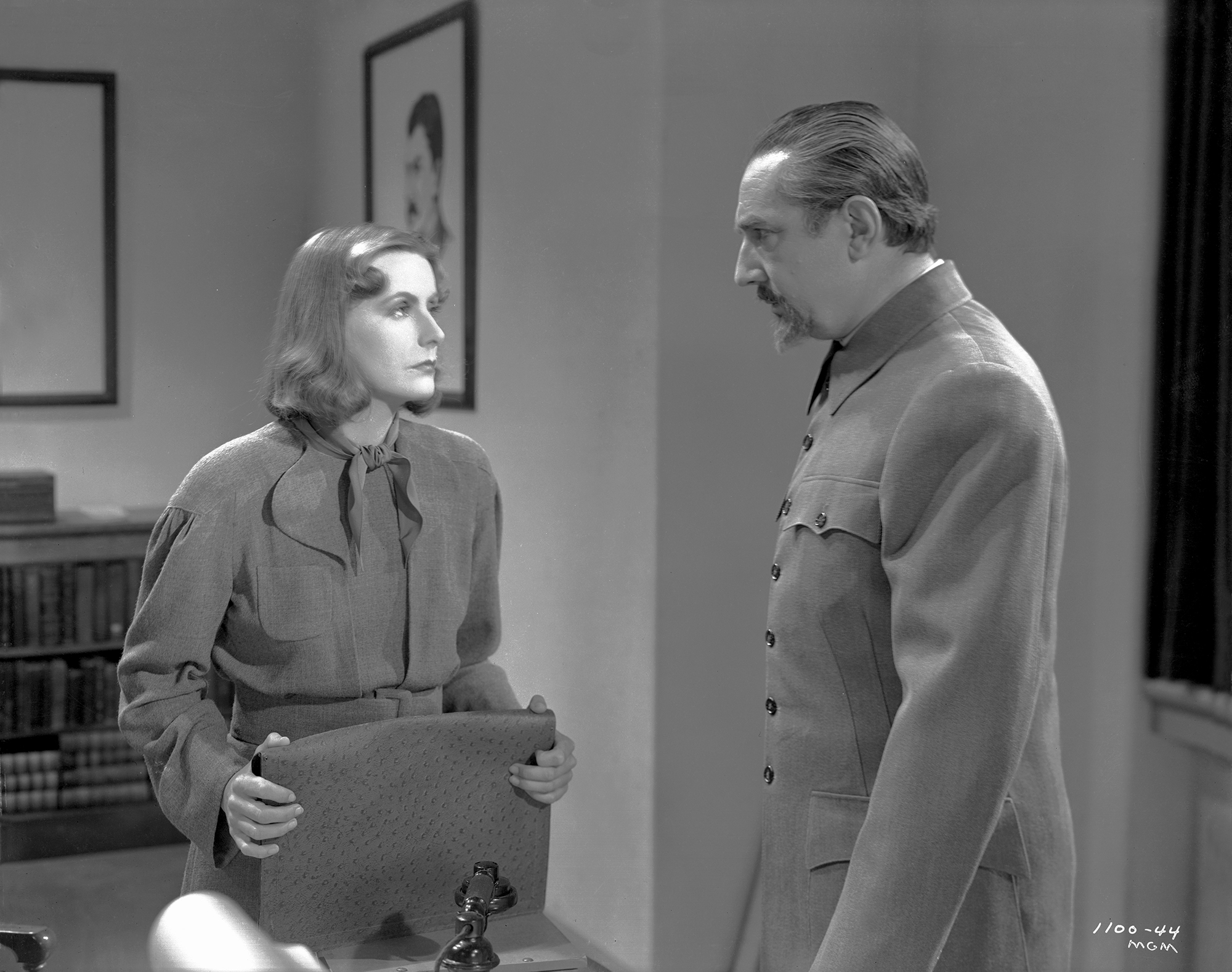 image from the 1939 movie "Ninotchka." Standing on the left is star Greta Garbo as the title character. She is looking at and speaking with the character in front of her, on the photo's right, played by Bela Lugosi, who has a mustache and goatee. Both characters are wearing gray attire.