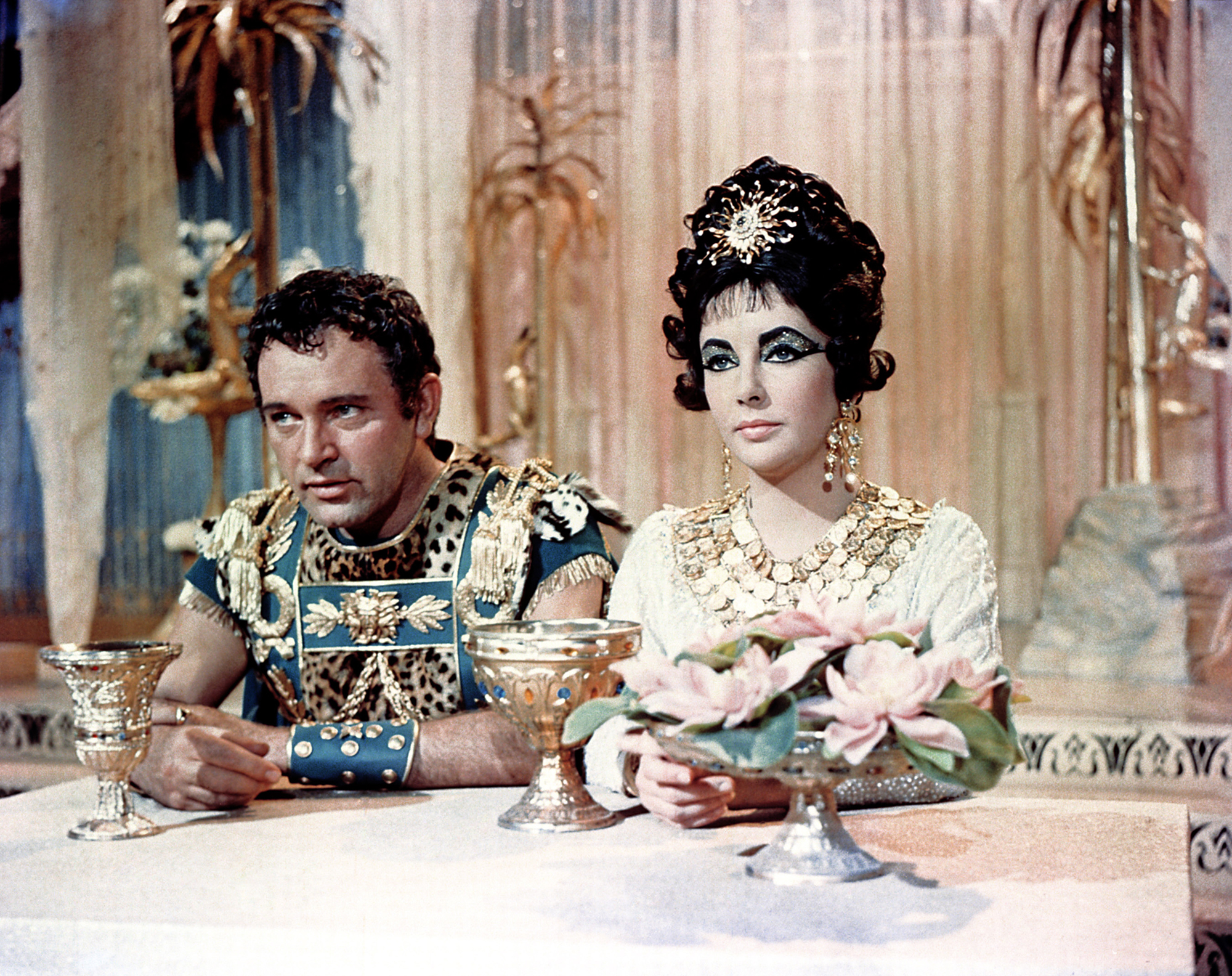 image from the 1963 movie "Cleopatra." Seated on the left at a table is Richard Burton as Mark Anthony. Seated directly next to him, on his left, is Elizabeth Taylor as Cleopatra.