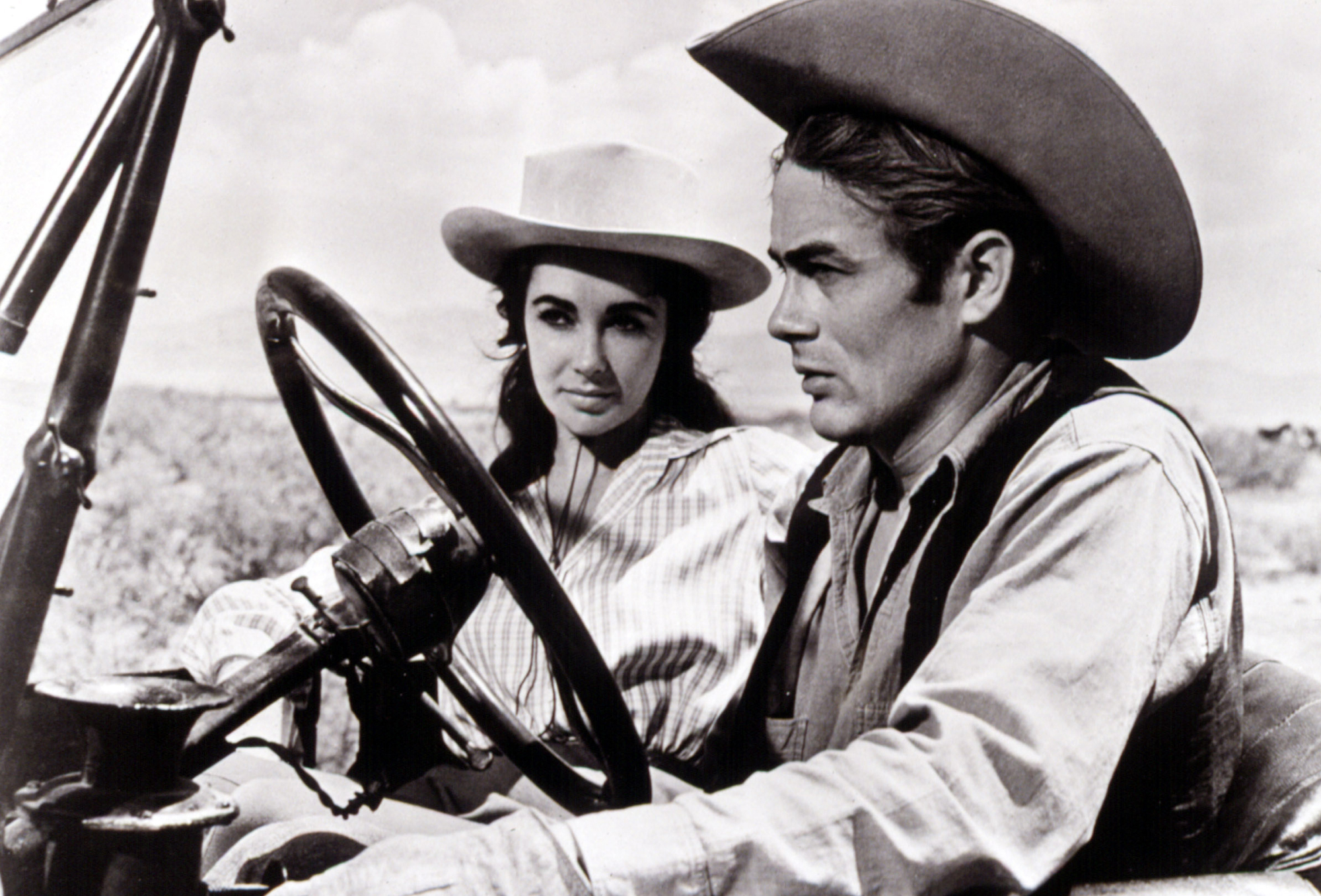 black and white image from the 1956 movie "Giant." it is a medium shot of stars James Dean, toward the front of the image and sitting behind the wheel of a car headed toward the left of the photo and wearing a cowboy hat, with costar Elizabeth Taylor in the passenger seat, wearing a white brimmed hat and looking at Dean as he looks ahead while driving.