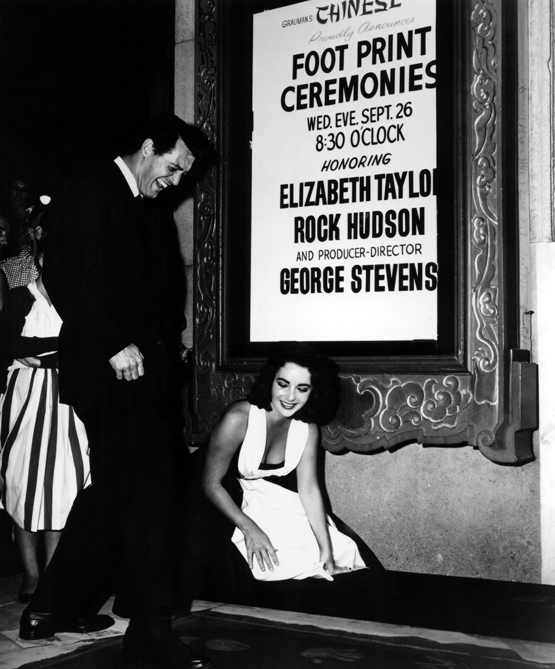 black and white image of Rock Hudson entering his footprints in the cement at Grauman's Chinese Theater at the premiere of the movie "Giant" in 1956. He is laughing as he steps into the cement, while costar Elizabeth Taylor, wearing a white dress, is kneeling down beside him and looking on.