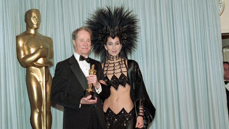 Don Ameche holding the Best Supporting Actor Oscar award for his role in Cocoon, backstage at the Academy Awards with award presenter Cher, who is wearing an unusual Bob Mackie evening gown with a feather headdress.