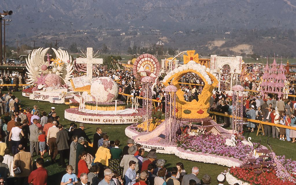 Panoramic view of prize-winning floats parked among throngs of visitors, at the public showcase following the 1956 Tournament of Roses Parade, in Pasadena, California, 1956. From left to right, are the City of Burbank Page One float, the Lutheran Layman's League Bringing Christ To The Nations float, and the Occidental Life Insurance Company of California's Rose Royalty Revisited float