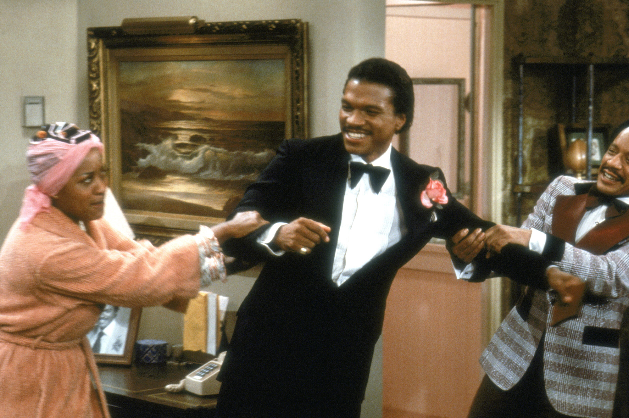 image from the 1978 "Me and Billy Dee" episode of "The Jeffersons". From left to right are Marla Gibbs as Florence, dressed in pink robe and curlers; Billy Dee Williams as himself, dressed in a standard black-and-white tuxedo; and Sherman Hemsley as George Jefferson, wearing a multicolored tux. Florence and George are having a tug-of-war over Billy Dee as the star smiles.