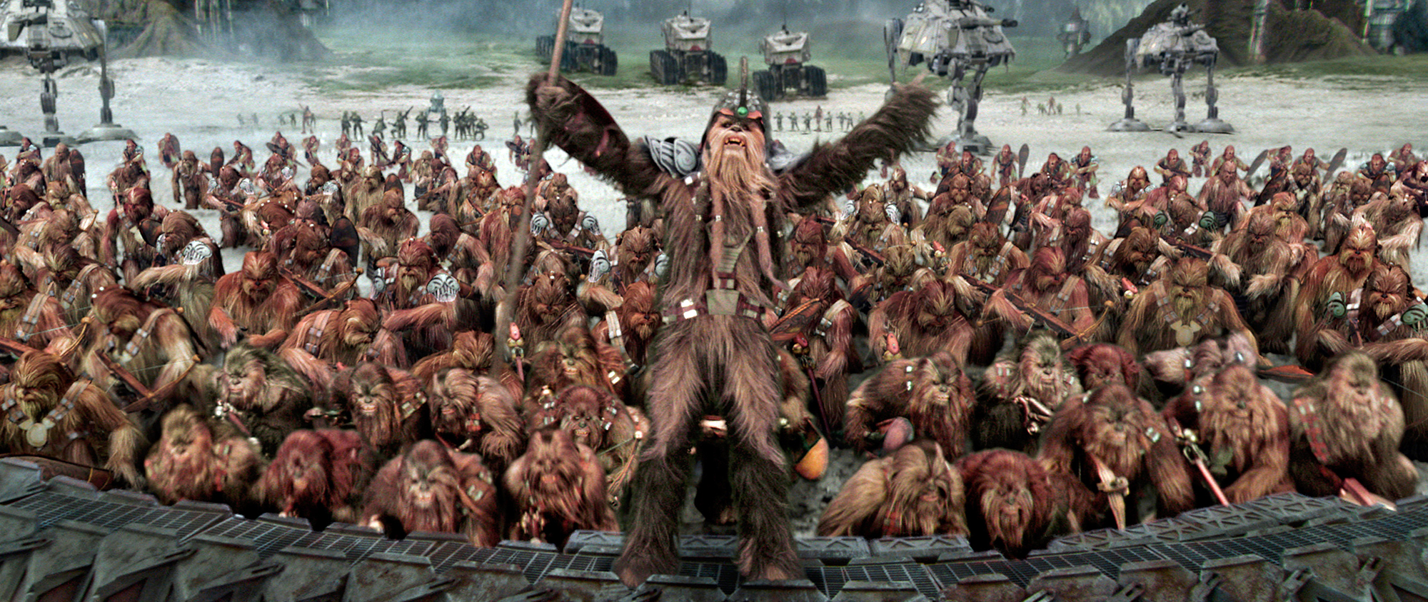 image from the 2005 movie "Star Wars: Episode III --- Revenge of the Sith." It is a long horizontal image of a scene on the Wookiees' home planet of Kashyyyk, with hundreds of the hairy humanoid beings gearing up for battle against an oncoming droid army. In the center, standing up above the rest of the Wookiee army, is a leader, wearing a helmet, holding a staff in his right arm and roaring.